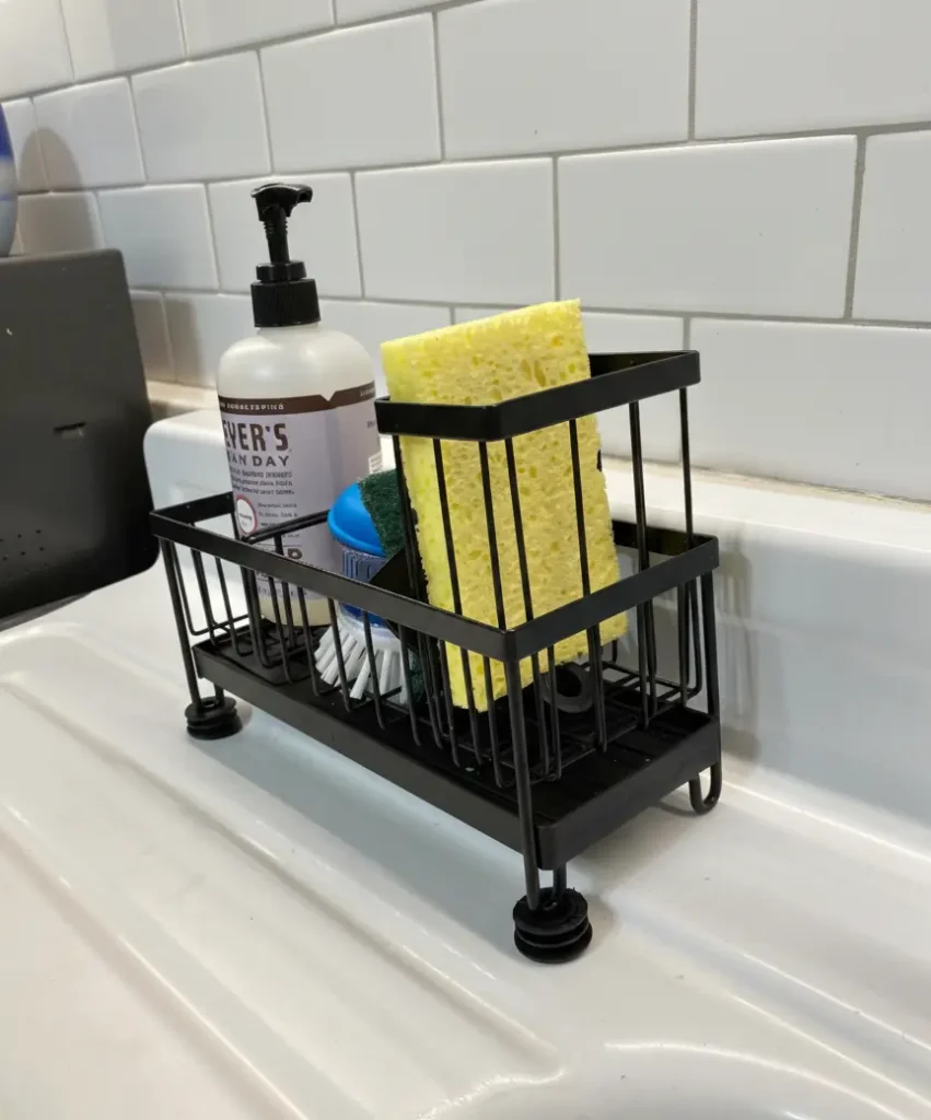 Cisily kitchen sink caddy organizer in black holding sponges, dish soap bottle, and brushes next to a stainless steel sink on a wooden countertop